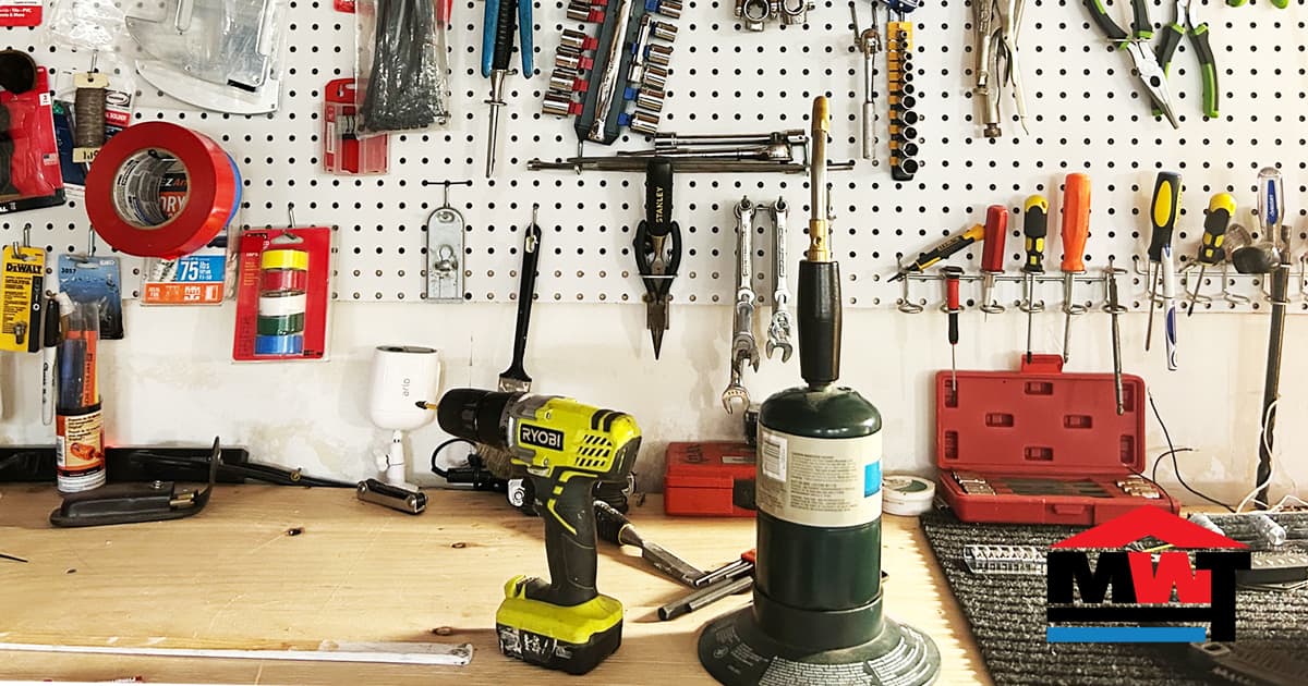 The Man With Tools workbench: pegboard of hand tools, a Ryobi drill and a torch, with the MWT logo